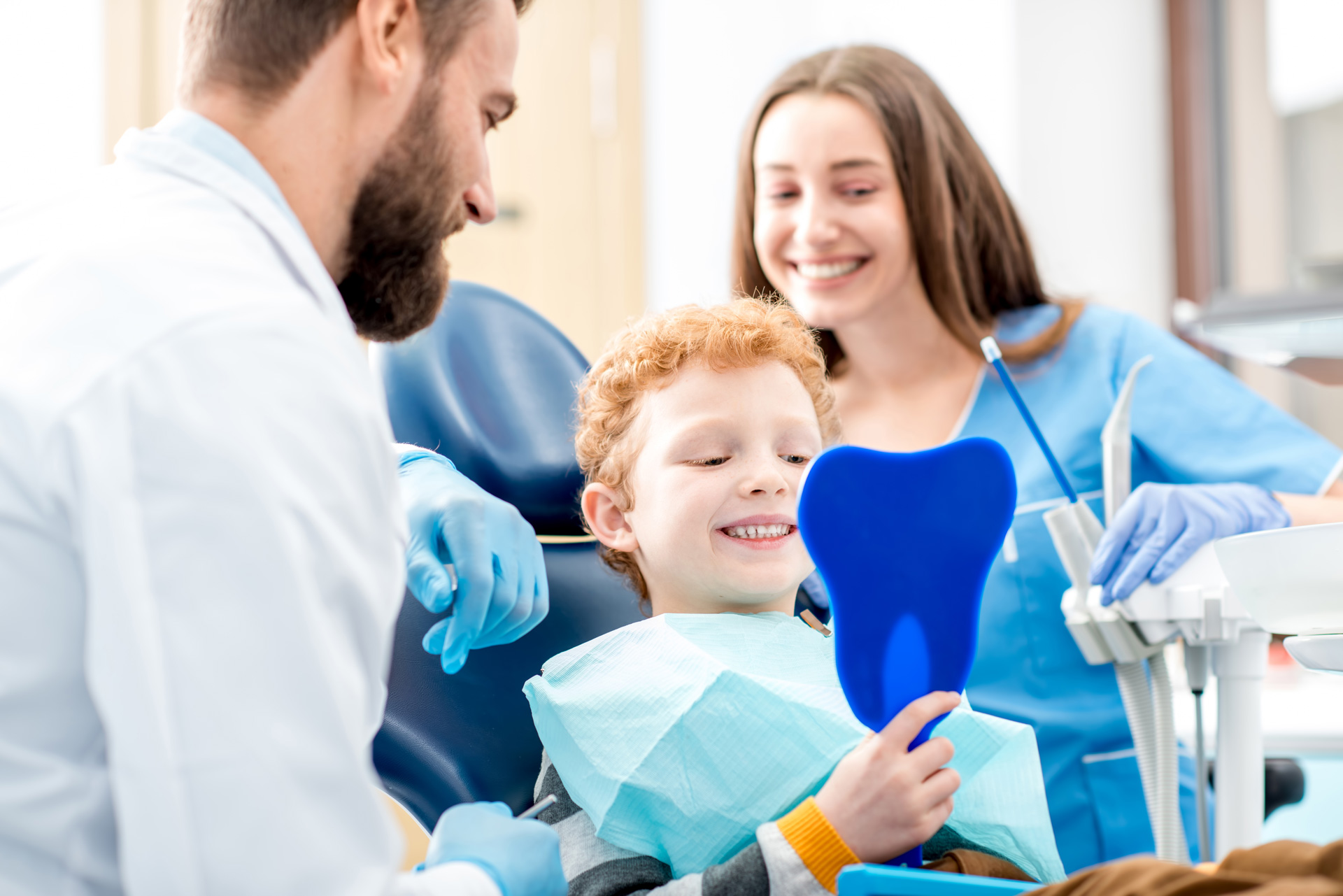 A smiling child holds a mirror while sitting in a dental chair.