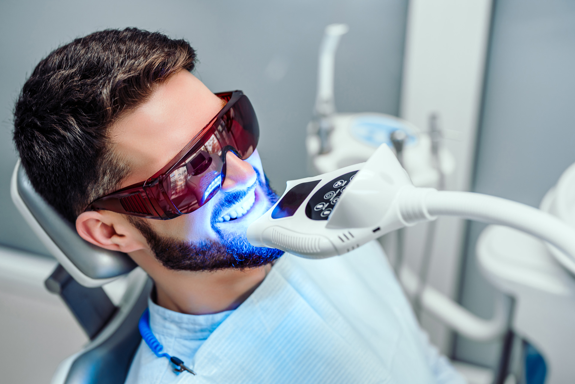 A man reclines in a dental chair, illuminated by a blue light focused on his teeth during a dental examination.