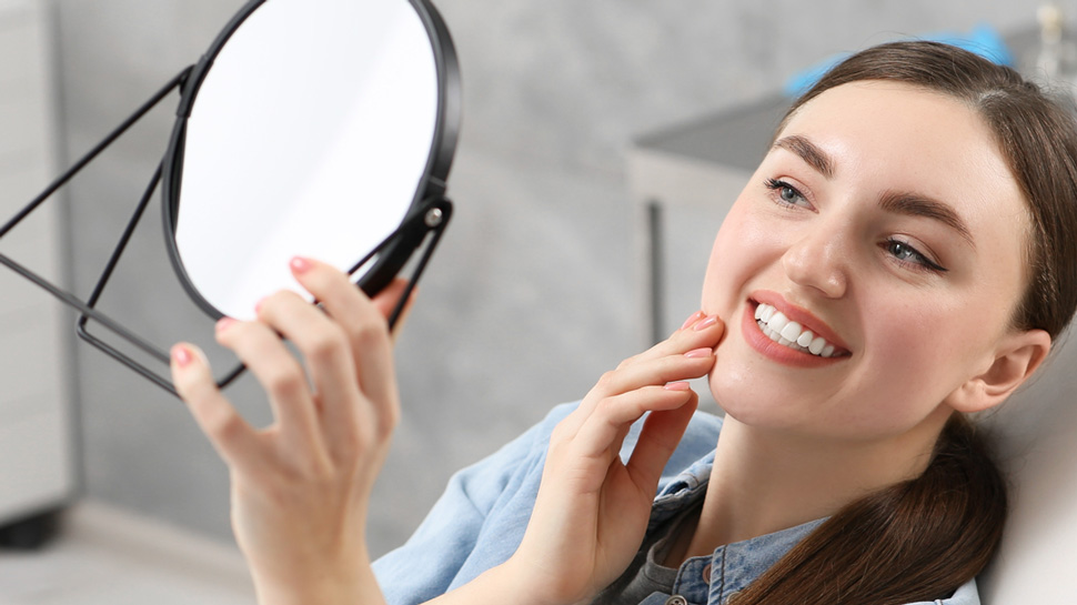 Person smiling while looking into a handheld mirror, touching their chin, against a blurred background.